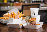 A close-up of a meal from the Better Burger Collection featuring golden fries in a white bowl, a small burger with lettuce on a brioche bun, and a white cup with a straw, all on a wooden table. The background is blurred, showing more food and drinks.