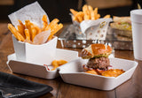 A white divided plate holds a burger with a pretzel bun, lettuce, and sauce, accompanied by tater tots. Next to it, a melamine fry box holds seasoned French fries. In the background, fries and a sandwich sit beside a white drink cup on a wooden table.