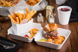 A tray holds a cheeseburger on a pretzel bun with lettuce, onion, and tomato, tater tots, pickle slices, and thick fries served in a dishwasher safe fry box, with ketchup on the side. A cup of purple drink and condiments sit on the wooden table.