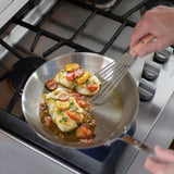 A person uses a Fish Turner with a wood handle to cook white fish fillets topped with sliced cherry tomatoes and herbs in a stainless steel pan on a gas stove. The fish sizzles in oil, and stovetop knobs are visible on the right side.