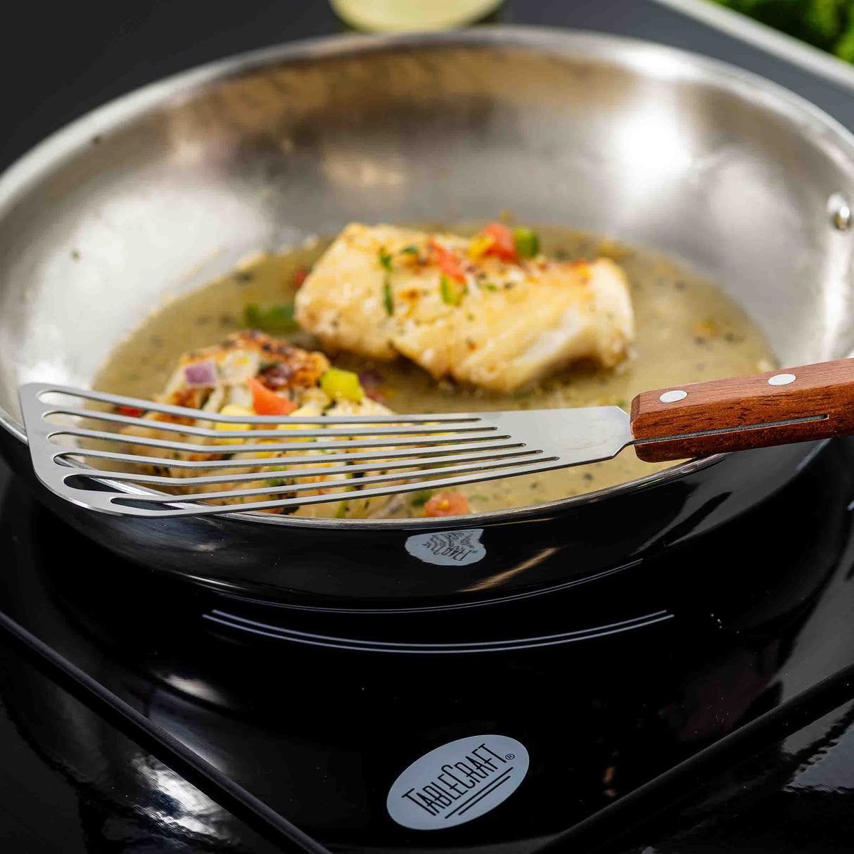 A fish fillet cooks in a stainless steel pan with sauce and herbs as a Fish Turner with a slotted blade and wood handle lifts or turns the fish. The pan sits on a black induction cooktop labeled “TableCraft.”.