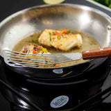 A fish fillet cooks in a stainless steel pan with sauce and herbs as a Fish Turner with a slotted blade and wood handle lifts or turns the fish. The pan sits on a black induction cooktop labeled “TableCraft.”.