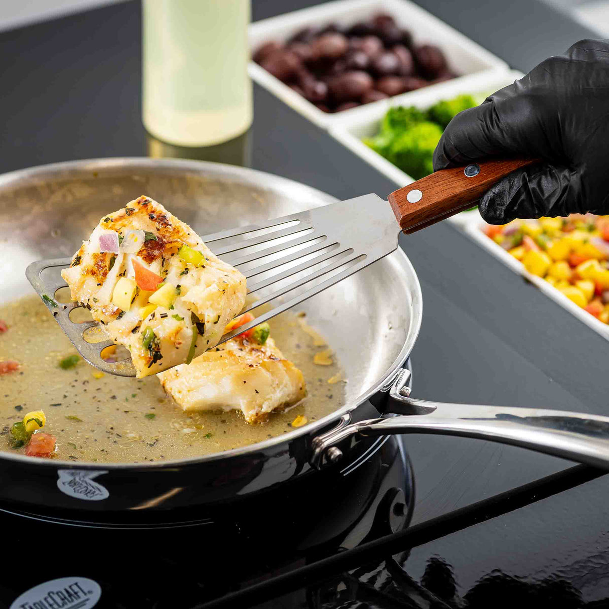 A gloved hand uses a Fish Turner with a slotted blade and wood handle to lift a cooked, seasoned fish fillet from a stainless steel pan with sauce. Colorful chopped vegetables and olives are arranged in dishes on a black countertop.