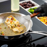 A gloved hand uses a Fish Turner with a slotted blade and wood handle to lift a cooked, seasoned fish fillet from a stainless steel pan with sauce. Colorful chopped vegetables and olives are arranged in dishes on a black countertop.
