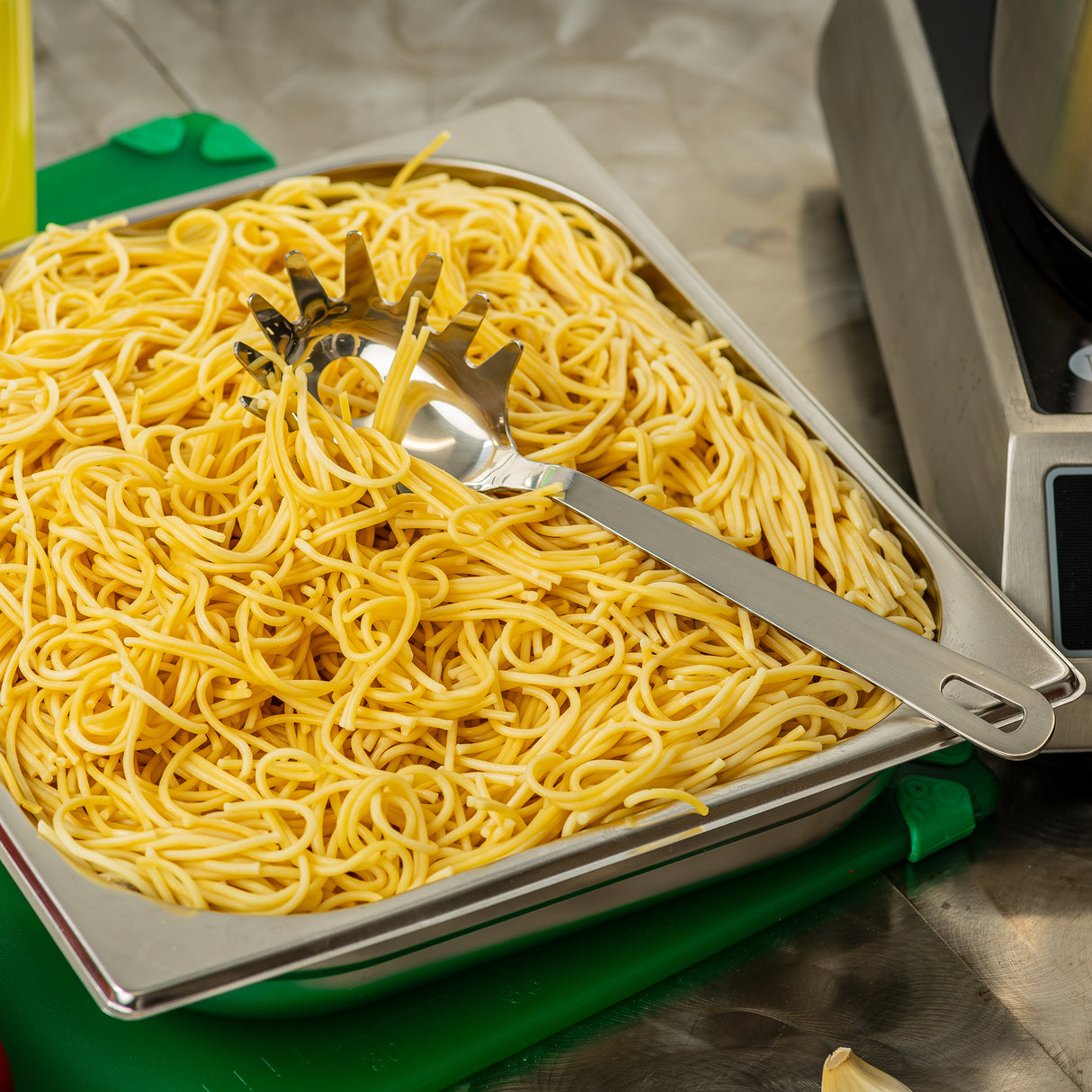 A metal tray filled with cooked yellow spaghetti noodles sits on a green cutting board. A stainless steel pasta grabber rests on top of the noodles. Part of a stove and other kitchen equipment are visible in the background.