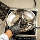 A person wearing a black glove uses a stainless steel Pasta Grabber to transfer cooked black pasta from a metal strainer into a frying pan on an electric stovetop. The stovetop display shows a temperature setting.