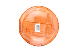 A round, mahogany wooden plate features a checkered pattern of varying wood grain shades, resembling woven wood. A white rectangular barcode sticker with black text sits in the center. The background is plain white.