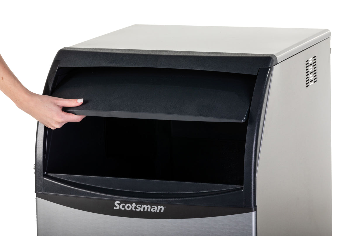 A person’s left hand lifts the black flap of a stainless steel Scotsman Ice Maker. The undercounter ice maker is mostly closed, with only the open hatch revealing its dark interior against a plain white background.