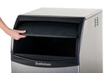 A left hand opens the black swing door of a stainless steel undercounter ice maker labeled Scotsman. The machine, photographed against a white background, reveals the interior where ice is stored. Only the hand and part of the arm are visible.