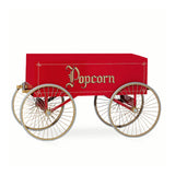 A vintage red Gay 90s Popcorn Wagon with four large gold-spoked wheels and “Popcorn” in ornate gold lettering on the side. The cart, by Gold Medal Products, features a rectangular body and a small wooden handle. The background is plain white.