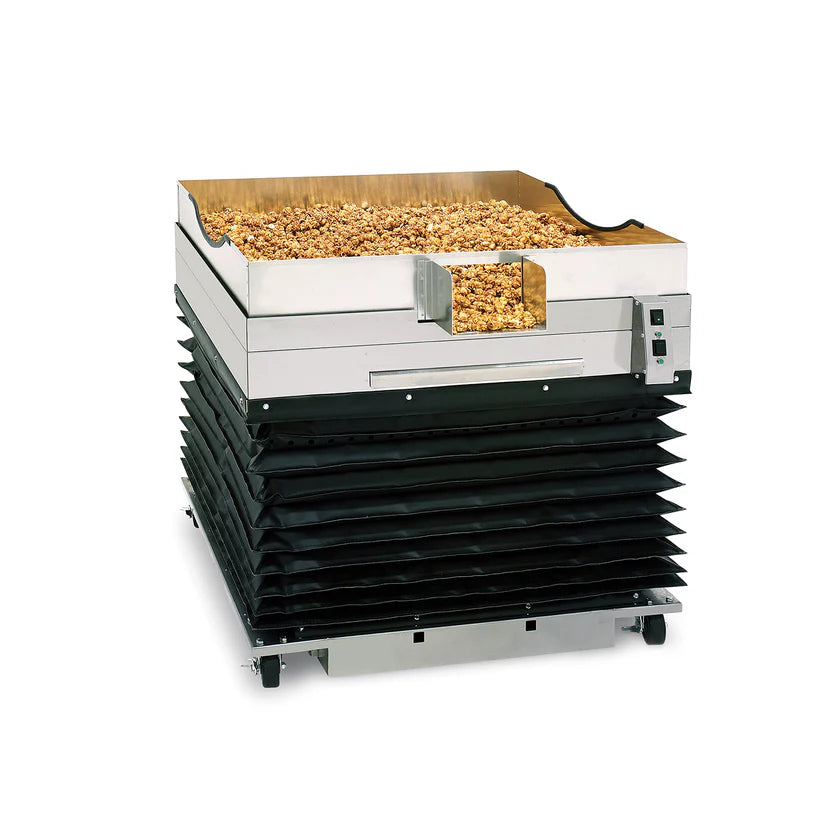 A large industrial food dehydrator with a metal tray filled with chopped nuts features a forced air cooling system, a silver and black exterior, digital control panel, vented accordion-like lower section, wheels for mobility, and sits against a plain white background.