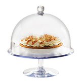 A glass cake stand with a clear polycarbonate dome cover displays a cream-topped pie. The pie has whipped topping and caramelized pieces on a golden crust, with the dome covering most of the pie. The background is plain white.