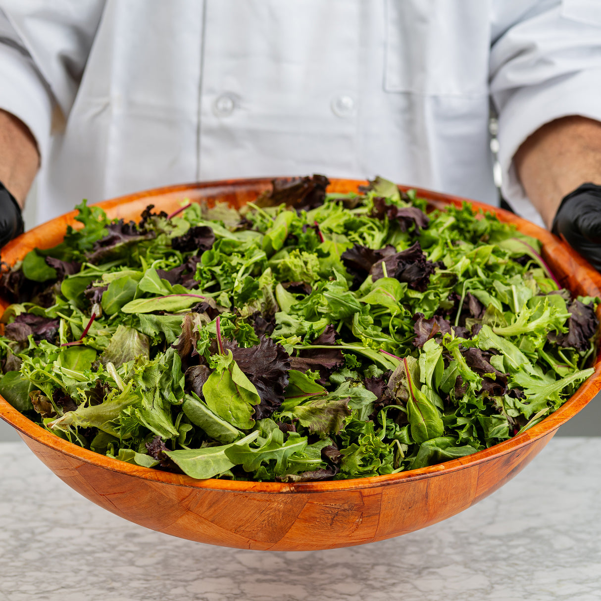 A person in a white chef’s coat and black gloves holds a mahogany salad bowl filled with mixed leafy greens. The bowl sits on a marble counter, showcasing vibrant green and purple lettuce leaves.