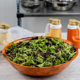 A large mahogany salad bowl filled with mixed leafy greens sits on a marble countertop. Three bottles of salad dressing are in the background, with a metal shelf and silver mixing bowls further behind them.