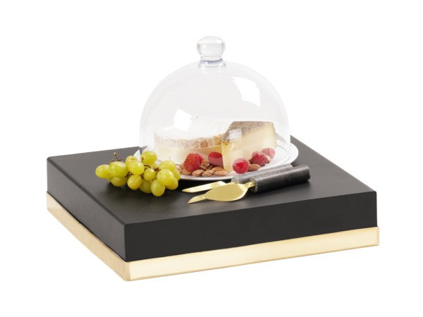 A glass cheese dome covers a wedge of cheese and berries on a clear plate. Next to it are green grapes, a cheese knife, and a spreader, all arranged on a black and gold Monaco-inspired serving tray with a sleek cube riser.