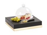 A glass cheese dome covers a wedge of cheese and berries on a clear plate. Next to it are green grapes, a cheese knife, and a spreader, all arranged on a black and gold Monaco-inspired serving tray with a sleek cube riser.