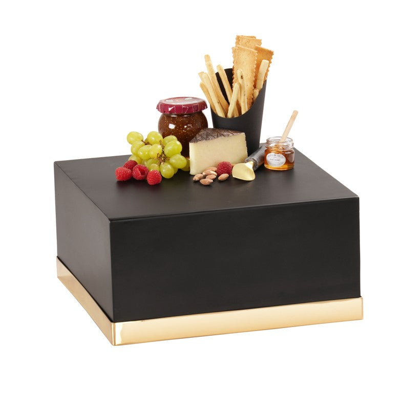 A black Monaco cube with a gold riser displays an assortment of snacks: green grapes, raspberries, cheeses, almonds, a small jar of honey with dipper, preserves, and a cup filled with breadsticks arranged on top.