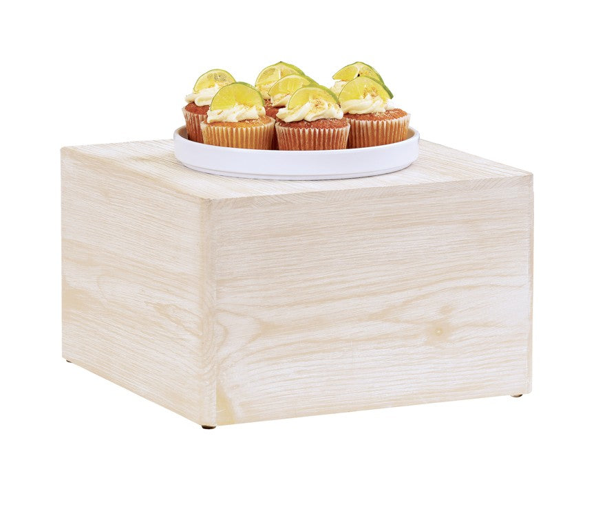 A square riser of white-washed pine wood holds a white round plate with six cupcakes, each topped with white frosting and a lime wedge. The background is plain white.