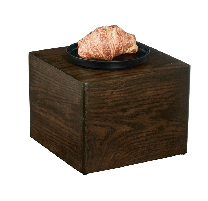 A golden-brown croissant sits on a round black plate, which rests atop a 12" x 12" x 9" Cal-Mil Heritage Riser with visible wood grain, set against a plain white background.