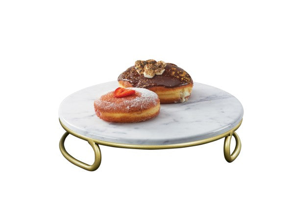 Two decorated donuts sit on a round, marble melamine cake stand with matte gold handles and legs. One donut is topped with chocolate and nuts; the other is coated in sugar with a red fruit garnish. The background is plain white.
