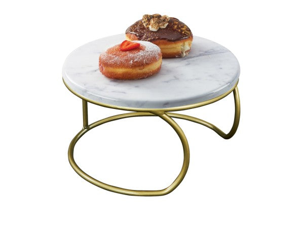 A small round table with a marble melamine top and a matte gold metal base holds two donuts, one topped with powdered sugar and strawberry, the other with chocolate glaze and nuts. The background is plain white.