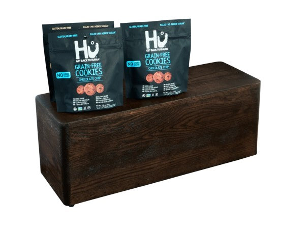Two black bags of Hu Grain-Free Chocolate Chip Cookies stand on a dark stained oak wood block. The bags feature images of cookies and bold white and blue text, highlighting that they are gluten-free, grain-free, and contain no added sugar.