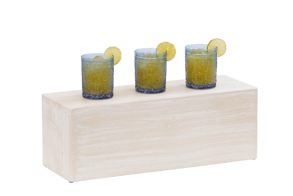 Three textured blue glasses filled with a yellow-orange drink, each garnished with a slice of lime, are arranged in a row on a rectangular pine wood riser against a white background.