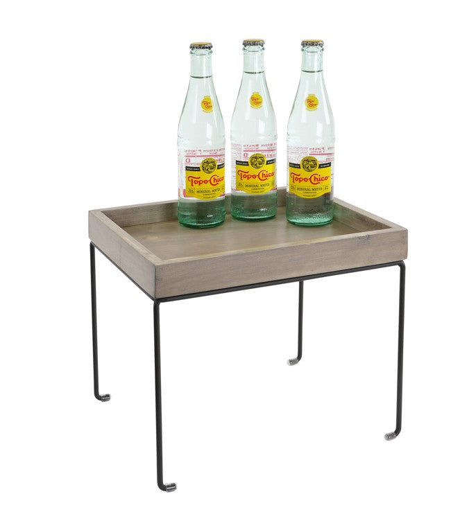 A small rectangular wooden tray table with black metal wire frame legs, similar to a Cal-Mil Aspen Riser, holds three glass bottles of Topo Chico mineral water with yellow labels and metal caps, neatly arranged side by side. The background is plain white.