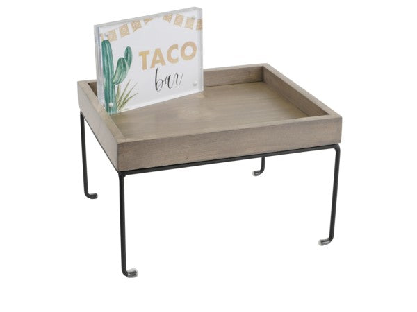 A small, square wooden Aspen Riser tray with black metal wire frame legs stands elevated. Inside the tray, an acrylic sign with a cactus and bunting reads TACO bar in green and gold letters. The tray features simple, modern styling and a rustic finish.