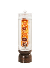 A tall, clear Heritage Beverage Dispenser with a spigot and a sturdy oak base, filled with water and slices of orange, lemon, and blood orange. The citrus slices are stacked vertically inside, gently infusing the water with fresh flavor.
