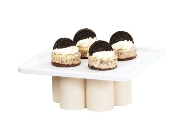 Four mini Oreo cheesecakes with cream topping and an Oreo cookie stand on a modern white cake stand with cylindrical legs and a pine wood riser base, all set against a plain white background.