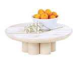 A round, white-washed pine wood coffee table with six cylindrical legs holds a white bowl with oranges and a branch of white flowers. The Newport-inspired design and clean riser detail create a minimal, modern aesthetic against the plain white background.