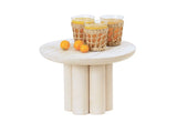 A small, round, white-washed pine wood table with a thick pedestal riser base holds three glasses of orange juice in wicker holders and two small oranges, all set against a plain white background.