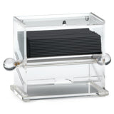 A clear acrylic stir stick dispenser holds a stack of black coffee stirrers. The box features rounded, clear knobs on both sides and an opening at the bottom front for easy access. Its modern, minimalist design complements any space.