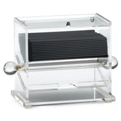 A clear acrylic stir stick dispenser holds a stack of black coffee stirrers. The box features rounded, clear knobs on both sides and an opening at the bottom front for easy access. Its modern, minimalist design complements any space.