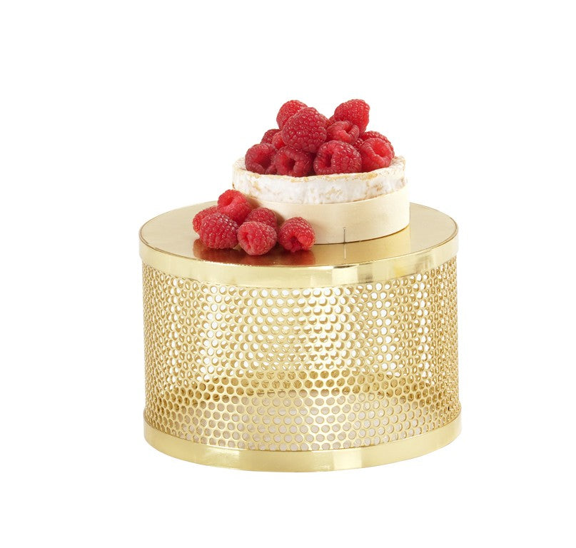 A round cheese wheel topped with fresh raspberries sits on a shiny gold riser inspired by Monaco, featuring a reflective surface and small, evenly spaced holes along its sides. The background remains plain white.