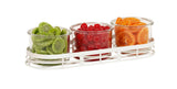 Three round glass jars in a white metal tray hold green candied slices, glossy red cherries, and orange citrus rounds, arranged from left to right for a vibrant condiment jar display against a white background.