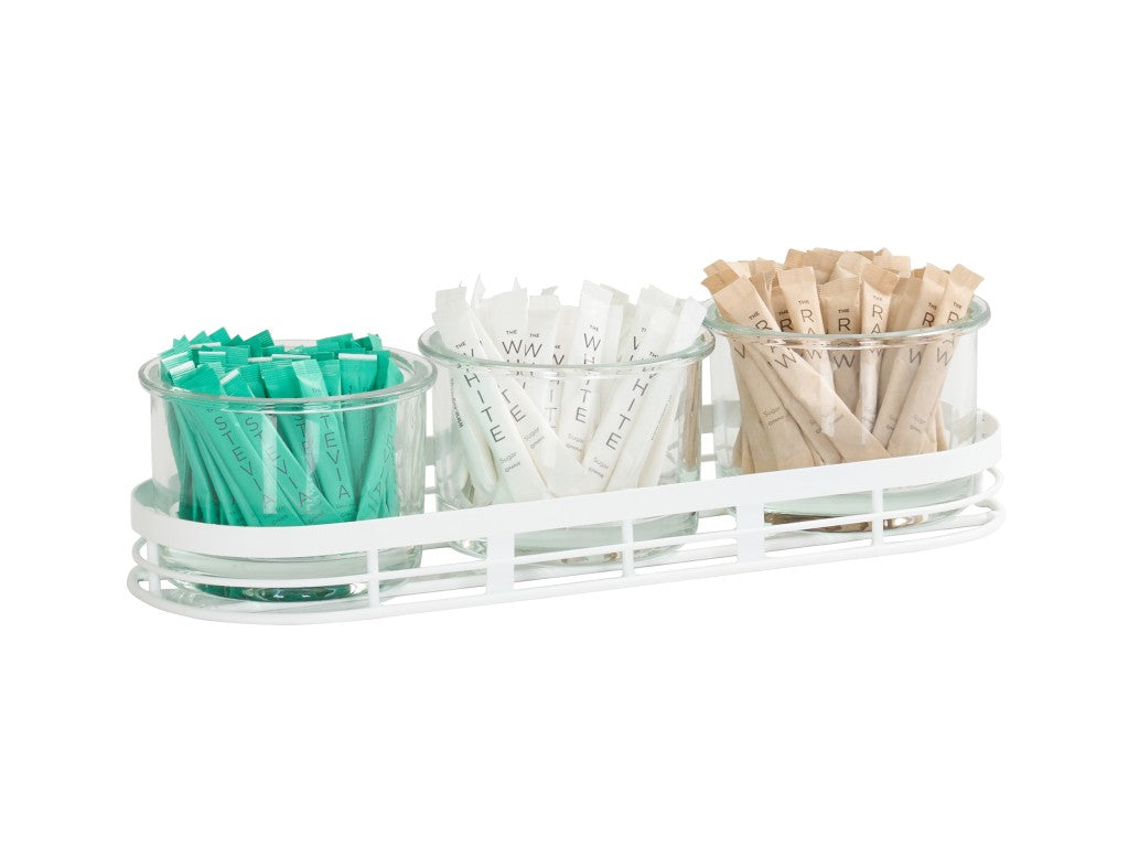 Three glass jars in a white metal tray create a tidy condiment jar display, holding teal, white, and brown sugar or sweetener packets neatly upright and grouped by color. The clear jars keep everything organized and easy to see.