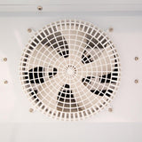 A close-up of a circular ventilation fan with black blades, covered by a white plastic grid. Mounted on a smooth white surface with visible screws, this fan is commonly found in Empura Refrigeration units like a glass door cooler. Photo taken head-on.