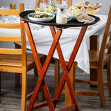 A wooden tray stand with a mahogany finish holds a black serving tray with salad dishes, dressing, and a napkin. Behind it, a dining table set with plates and bread is surrounded by wooden chairs for a welcoming meal.