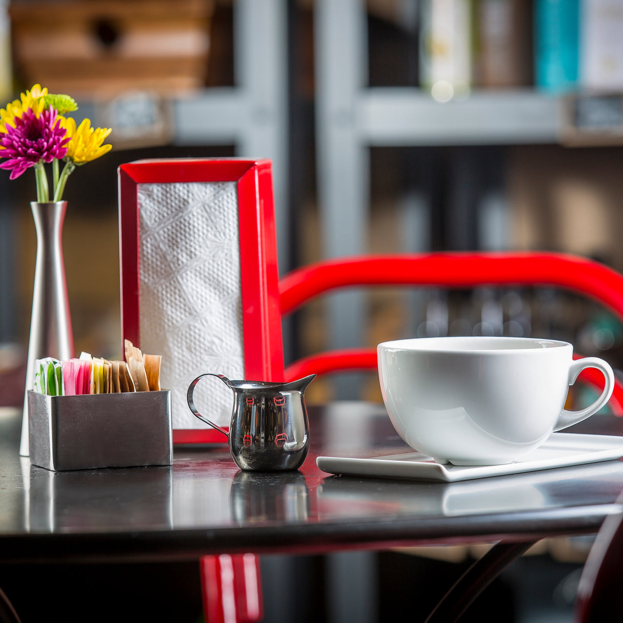 A white cup and saucer sit on a table beside a 3 oz stainless steel bell creamer, packets of sugar, a silver vase with colorful flowers, and a red napkin holder. A red chair and shelves are visible blurred in the background.
