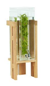 A modern, minimalist beverage dispenser features clear glass, a wooden stand, and a silver removable spigot. Filled with water and leafy herbs, its sleek design highlights natural wood and glass elements for stylish drink service.