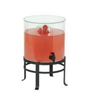 A clear cylindrical polycarbonate beverage dispenser with an infusion chamber holds pink liquid and floating strawberries, set on a black metal stand. The dispenser features a black spigot at the front for easy pouring.
