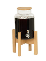 A clear glass drink dispenser filled with a dark liquid, likely coffee or tea, sits on a light wooden stand with four legs. Perfect for beverage display, it features a wooden lid and silver spigot. The background is plain white.