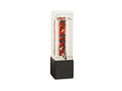 A tall, rectangular clear beverage dispenser with a black metal base features an infusion chamber filled with water and visible pieces of red, black, and white fruit. The spigot near the bottom allows easy pouring against a plain white background.
