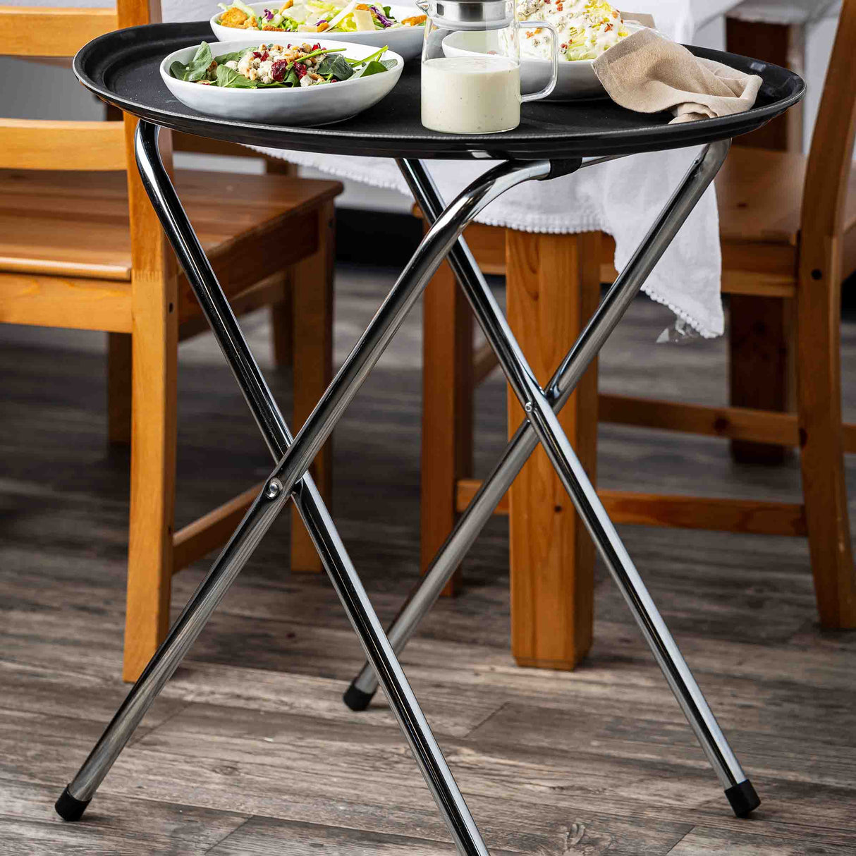 A round black serving tray rests on a chrome plated tray stand with tubular construction next to a wooden dining table and chairs. The tray holds plates of salad, a glass pitcher, a white gravy boat, and a beige napkin on the wood-look floor indoors.