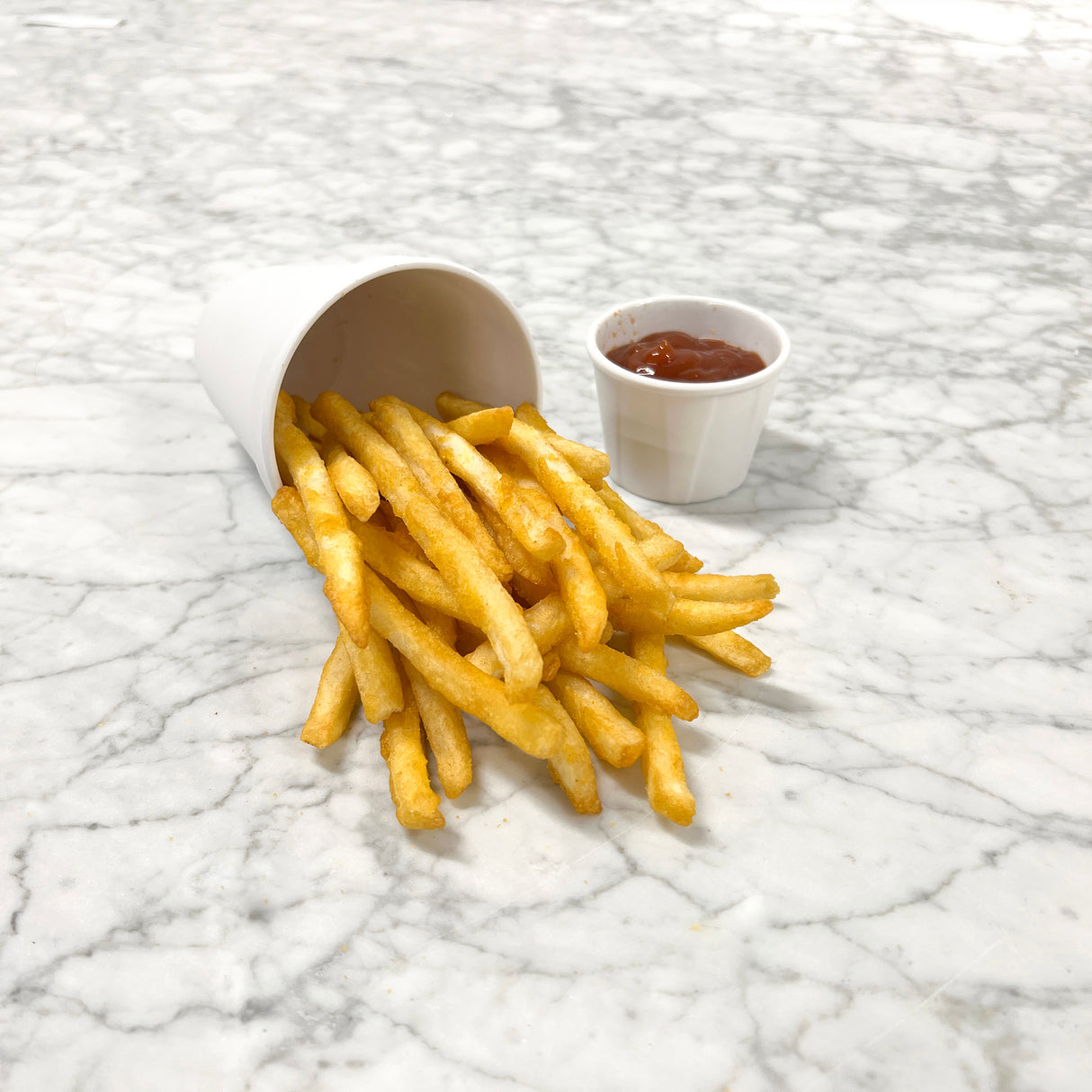 A white cup lies on its side, spilling golden French fries across a white marble surface. Next to the fries is a small melamine soufflé cup filled with red ketchup. The scene evokes the style of the Better Burger Collection.