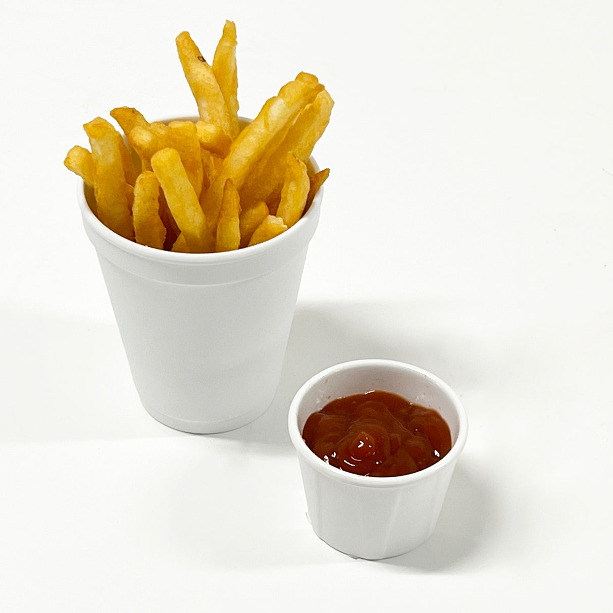 A white soufflé cup filled with golden French fries sits beside a small melamine container of ketchup, all placed on a plain white surface—part of the Better Burger Collection.
