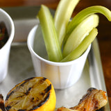 A small white soufflé cup holds several celery sticks. Next to it, there’s a grilled half lemon with char marks and a piece of cooked chicken, all on a metal tray. Part of another dish in a white bowl is visible at the edge.