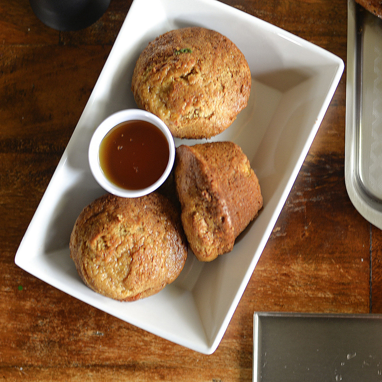 A rectangular white plate holds three golden-brown muffins and a small melamine soufflé cup of honey. The plate sits on a rustic wooden surface, with part of a metal tray visible in the bottom right corner.